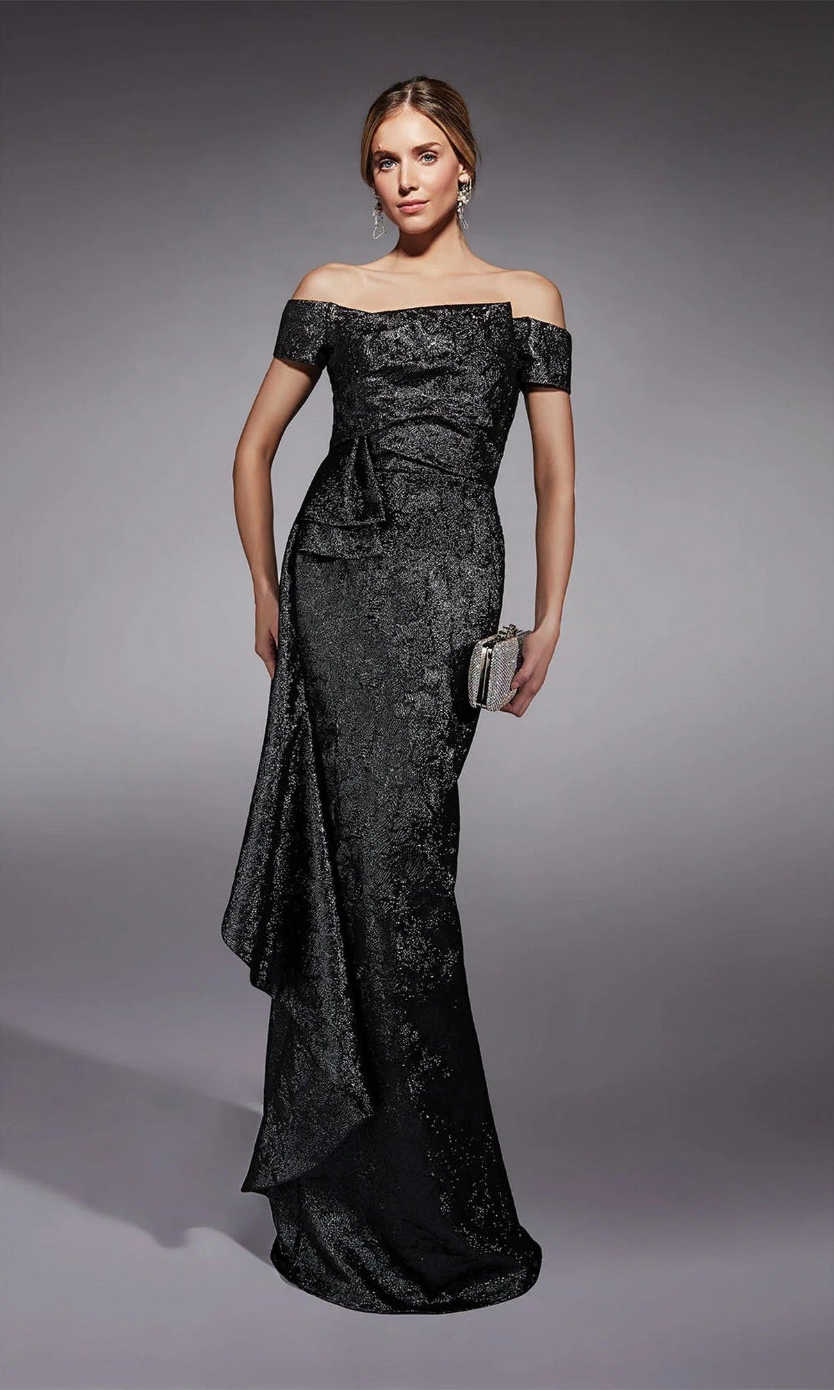 A woman stands against a plain gray background wearing the Alyce 27735, a textured black off-the-shoulder formal gown with side drape detail, holding a small clutch. Perfect elegance for the mother of the bride.